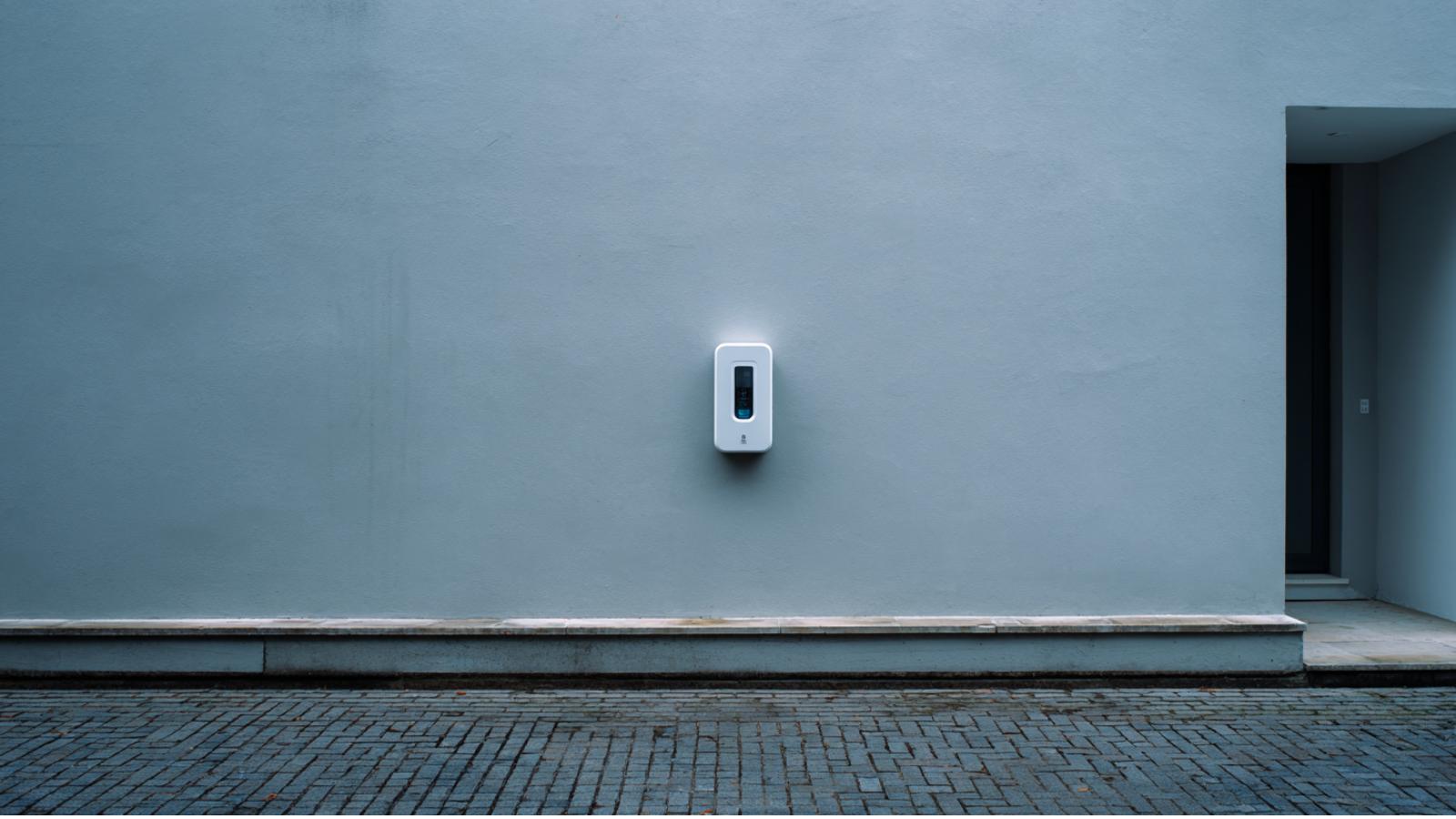 A white wall-mounted home EV charger on a pale rendered wall above a brick-paved drive